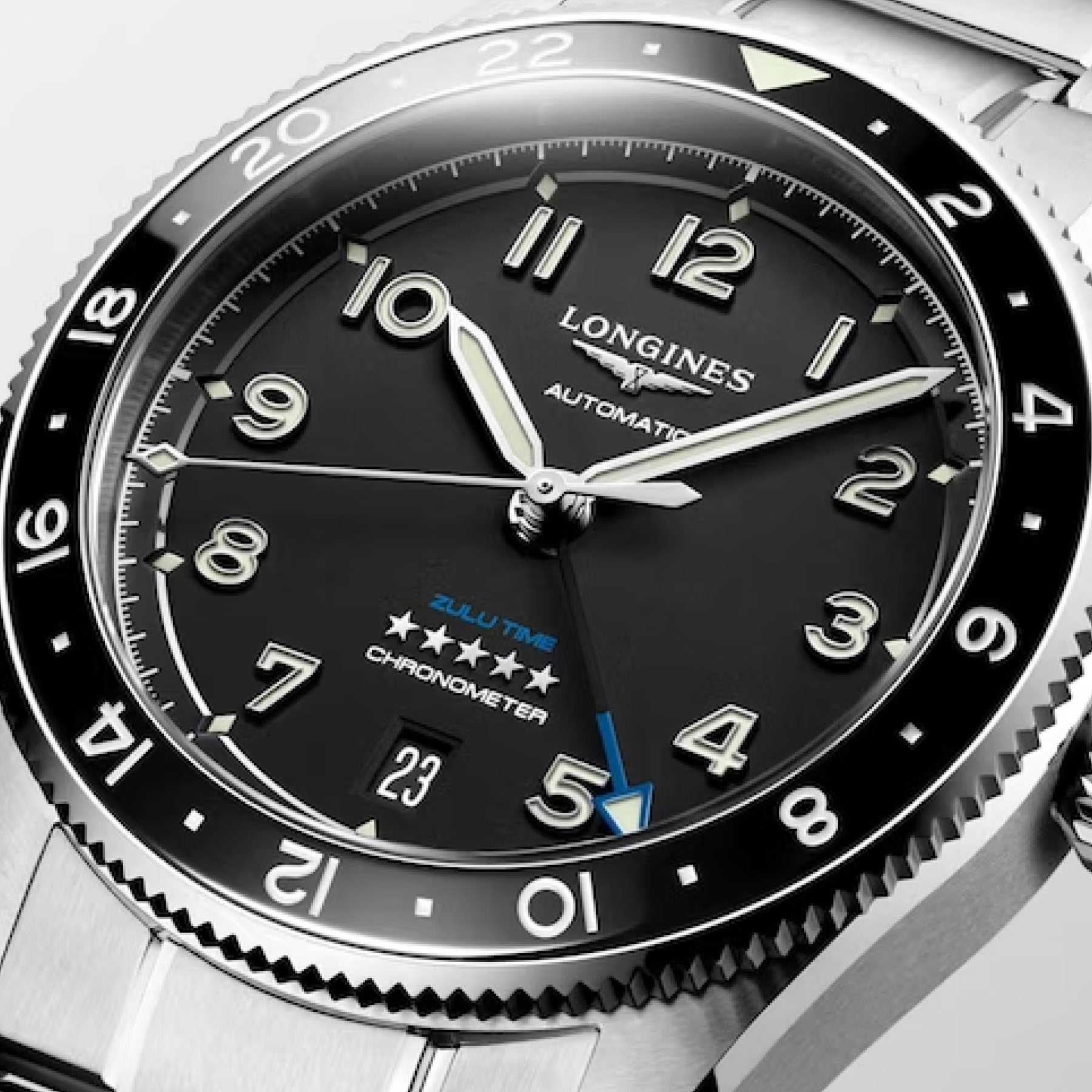 A close-up photo of the Longines Spirit Zulu Time Automatic COSC 39mm Watch highlights its sophisticated black dial with luminous Arabic numerals, silver hands, and a striking blue second time zone hand. Enhancing its elegance, it includes a date window at 6 o'clock next to the text "ZULU TIME CHRONOMETER.