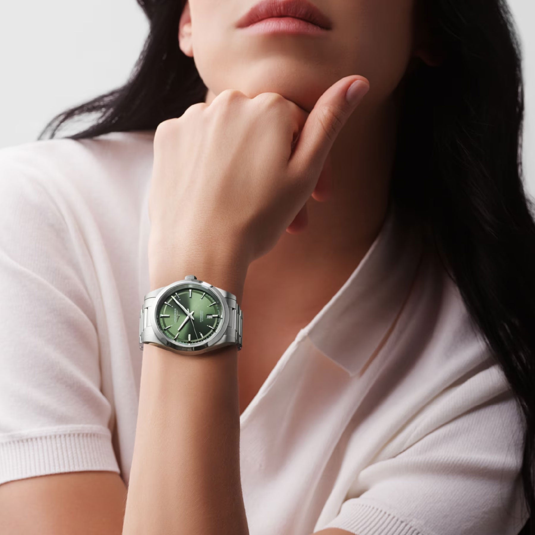 A person with long, dark hair, wearing a white collared shirt, rests their chin on their hand in a pose of quiet elegance. They are wearing the Longines Conquest Automatic 41mm Watch by Longines, featuring a green face. The background is a plain light gray, enhancing the scene's serene atmosphere.