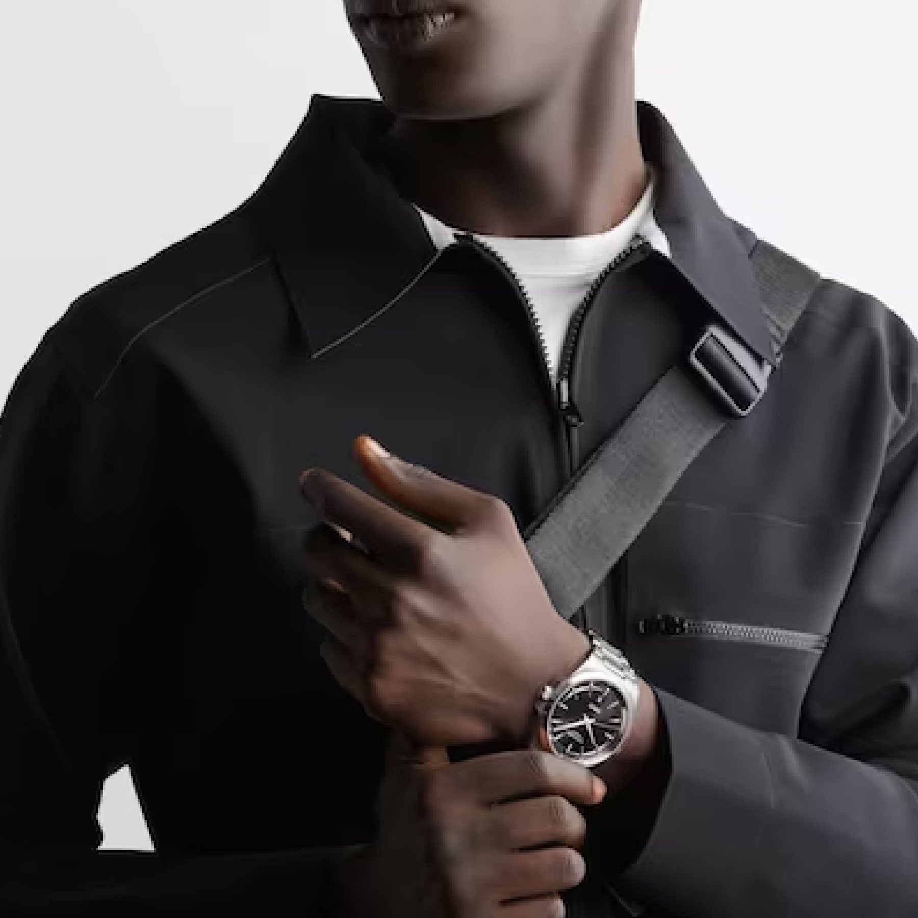 A person stands with arms crossed, dressed in black and showcasing a prominently visible Longines Conquest Automatic 41mm watch on their wrist. They have a crossbody strap over one shoulder against a plain light-colored background.