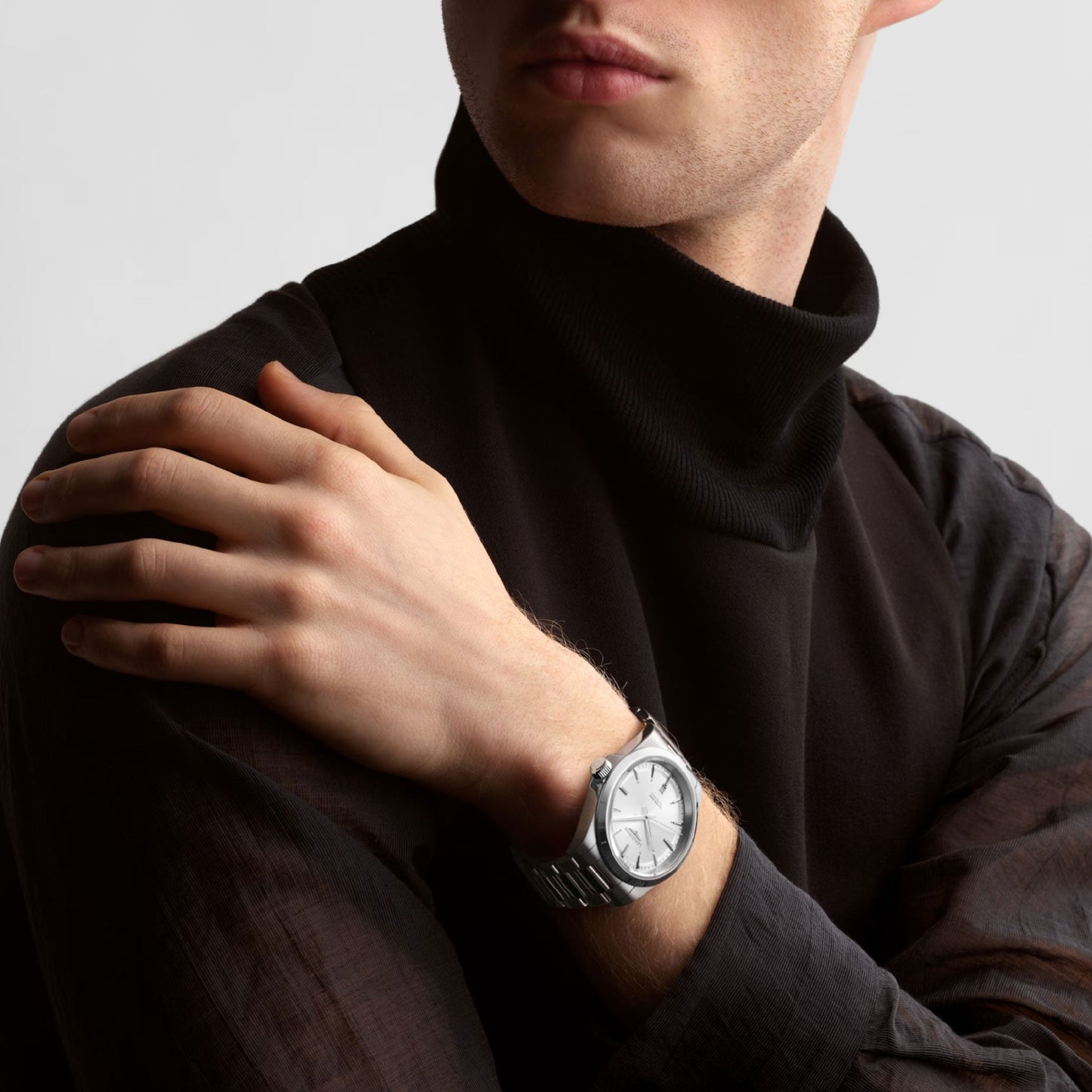 A person exudes elegance in a black turtleneck sweater and a Longines Conquest Automatic 41mm watch, featuring a silver band and white face. Their right hand rests gracefully on their left shoulder against a plain light background, capturing an understated sense of sophistication.