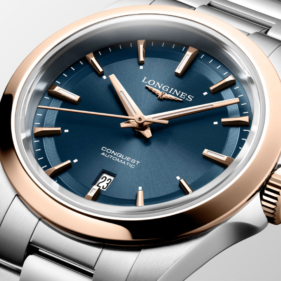A close-up of the Longines Conquest Automatic 30mm Watch by Longines, featuring a blue dial, gold-tone hands and markers, silver and gold bezel, date at 6 o’clock, and a stainless steel bracelet—sporty elegance for everyday wear.