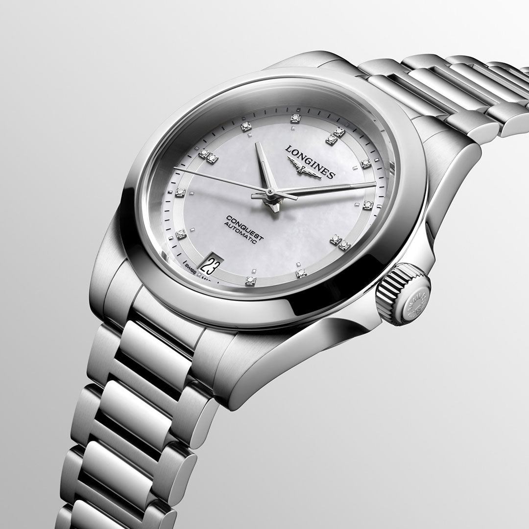 The Longines Conquest Automatic 34mm Watch exudes elegance with its silver-tone metal band and diamond hour markers. Featuring a date display at the 3 o'clock position, it showcases a white face with silvery hands on a light gray backdrop, offering both style and performance.