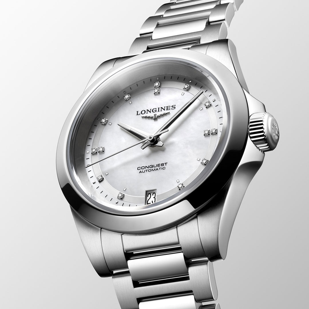 The Longines Conquest Automatic 34mm Watch epitomizes elegance with its pearl white dial and diamond hour markers. A date display at 6 o'clock perfectly complements the stainless steel bracelet, while its sleek design seamlessly blends performance and style against a light gray backdrop.