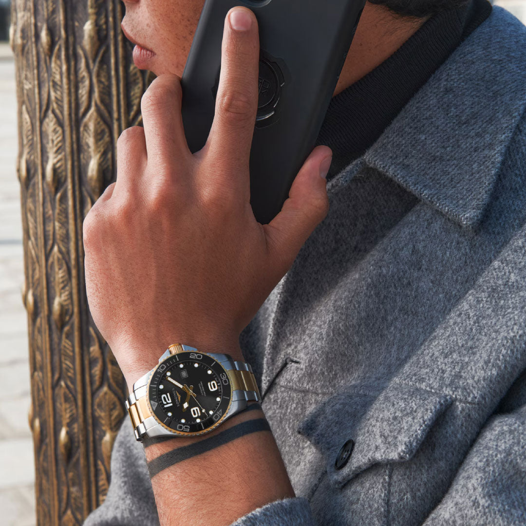 A person in a grey coat holds a smartphone to their ear, revealing a Longines Hydroconquest Automatic 43mm Watch with a black dial and gold and silver band on their left wrist. An ornate, textured pole appears in the background.