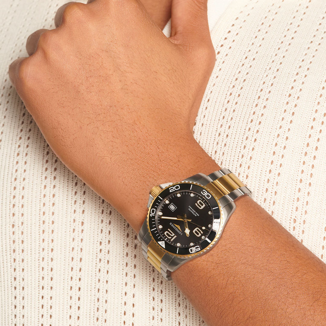 A close-up of a wrist wearing the Longines Hydroconquest Automatic 43mm Watch, featuring a two-tone gold and silver band, black face, and white markings, set against textured white knit fabric.