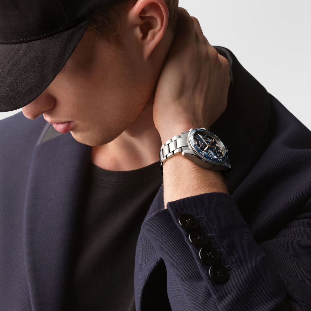 A person wearing a black cap and dark blazer looks down, touching their neck. Adorning their wrist is the Longines Spirit Flyback Automatic Chronograph 42mm watch, embodying the sophistication of the Longines brand. The background is plain and light-colored.