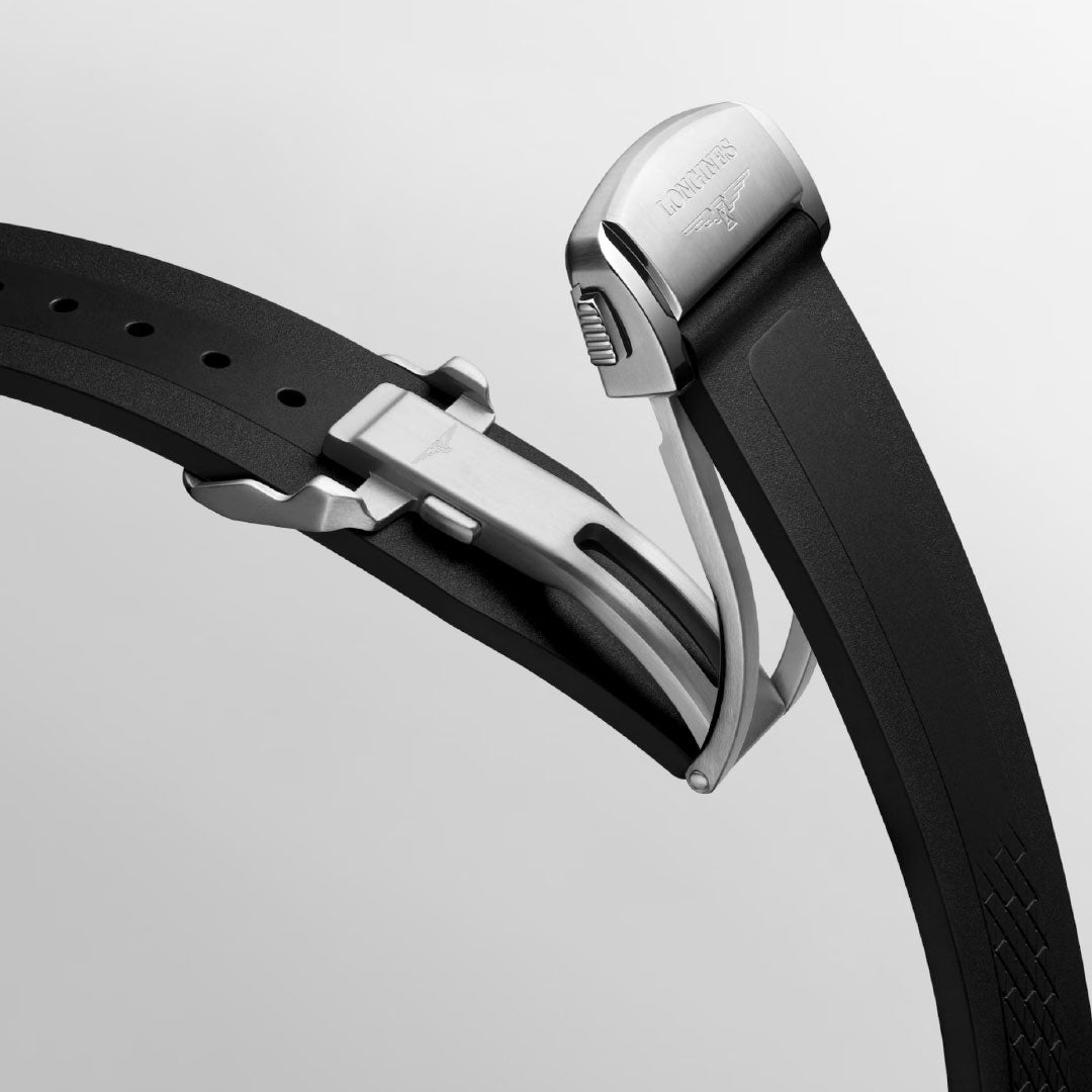 A close-up of the Longines Conquest Chronograph Automatic 42mm watch shows a black strap with a stainless steel clasp engraved with the Longines logo, featuring a self-winding mechanical movement against a light gray background.