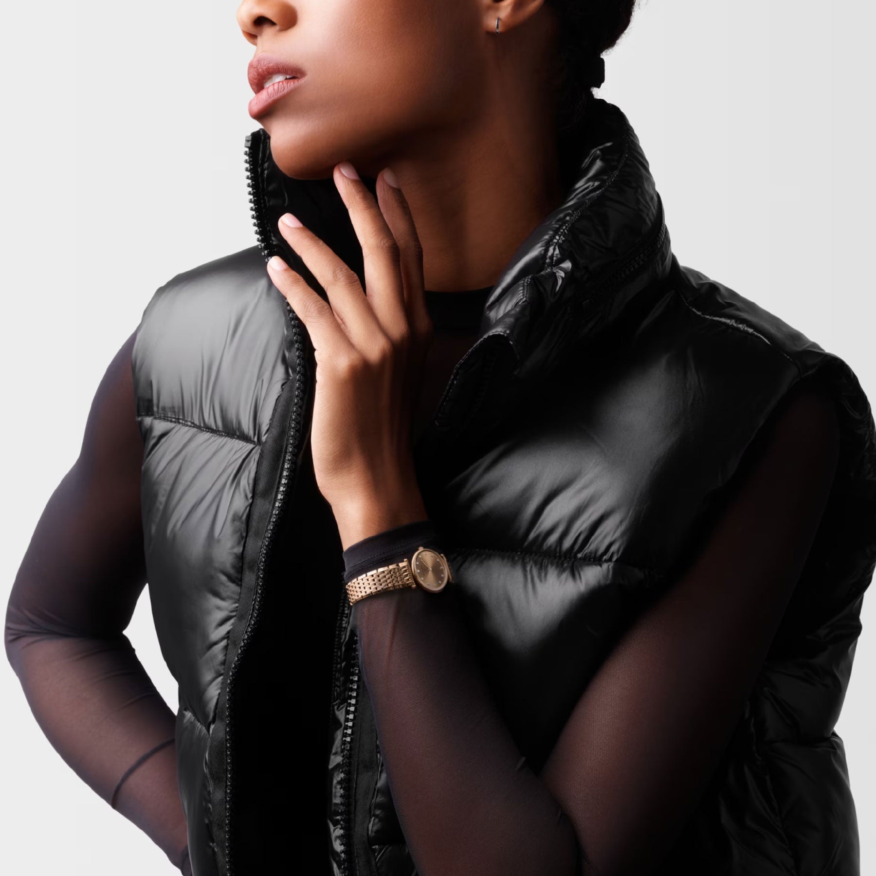 Dressed in a shiny black puffer vest layered over a sheer long-sleeve top, a person emits an air of timeless sophistication with one hand thoughtfully touching their chin. They elegantly wear the Longines La Grande Classique De Longines Quartz 24mm Watch, set against a neutral backdrop.