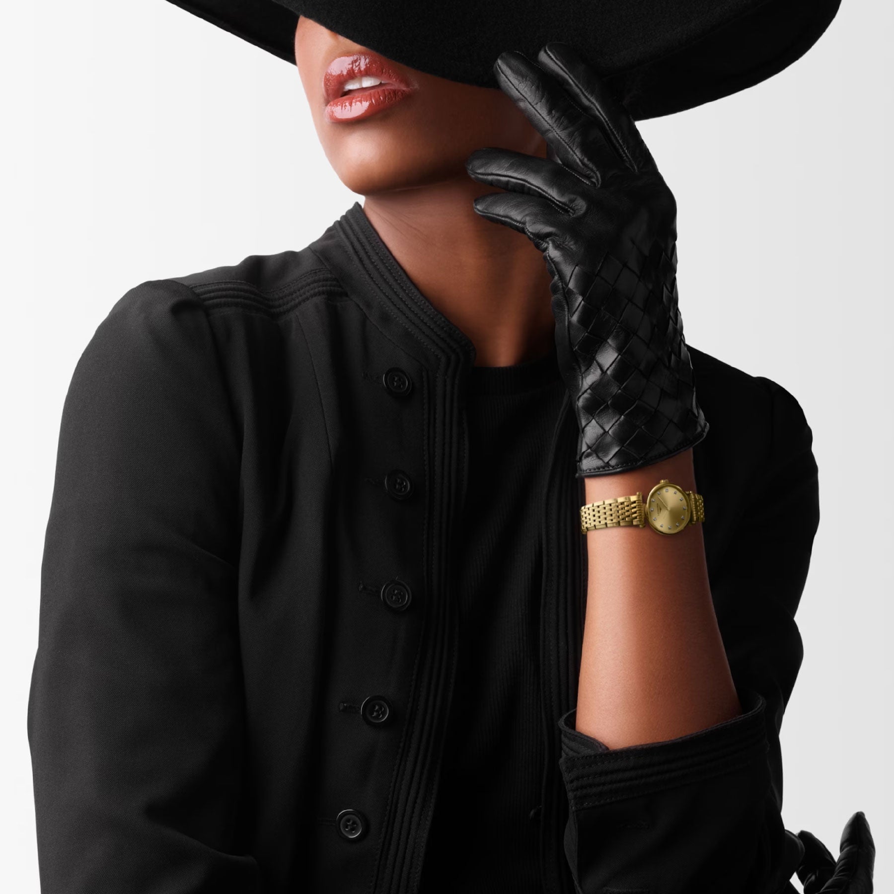 A woman dressed in black, adorned with textured gloves and a wide-brim hat casting a shadow over her face, embodies timeless sophistication. Her Longines La Grande Classique De Longines Quartz 24mm watch gleams subtly in gold. With lips painted for a pop of color, the image exudes classic elegance and mystery.