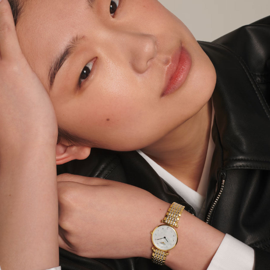 A person in a black leather jacket rests their head on one hand, showcasing a Longines La Grande Classique Quartz 24mm Watch with a mother-of-pearl dial on their wrist. The image highlights both the elegant Longines watch and their relaxed expression.