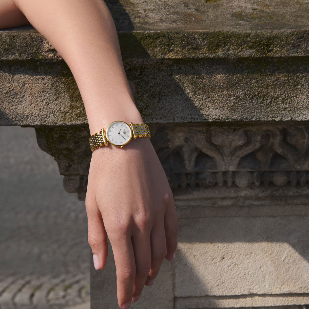 A person's hand wearing the Longines La Grande Classique Quartz 24mm gold wristwatch rests on an ornately carved stone ledge outdoors, highlighted by natural light.