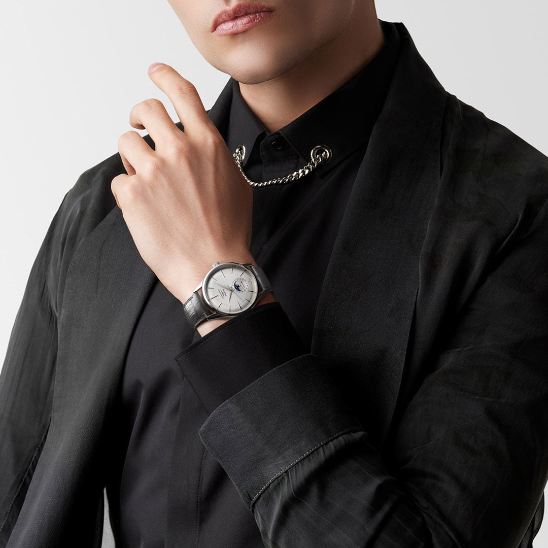 A person in a black blazer and shirt poses with their hand near their chin, showcasing the striking Longines Flagship Heritage Automatic 38.5mm Watch. A moon phase complication adds to its allure, while a chain necklace complements the look against the plain white background.