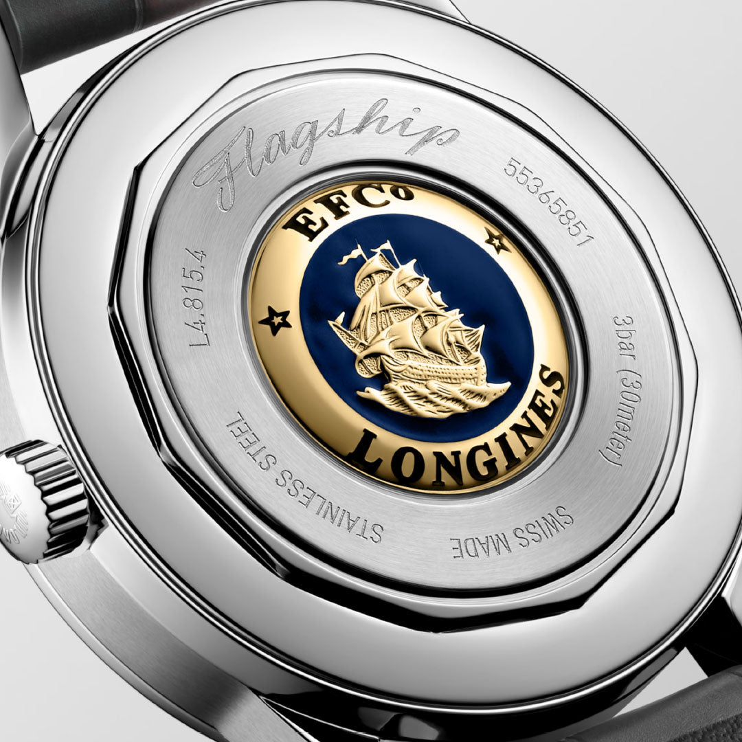 A close-up view of the Longines Flagship Heritage Automatic 38.5mm watch showcases a nautical-themed medallion with a ship illustration on its back. The encircling text includes "Flagship," the brand name "Longines," and details like "Stainless Steel," "Swiss Made," along with serial numbers, emphasizing the exclusive movement within.