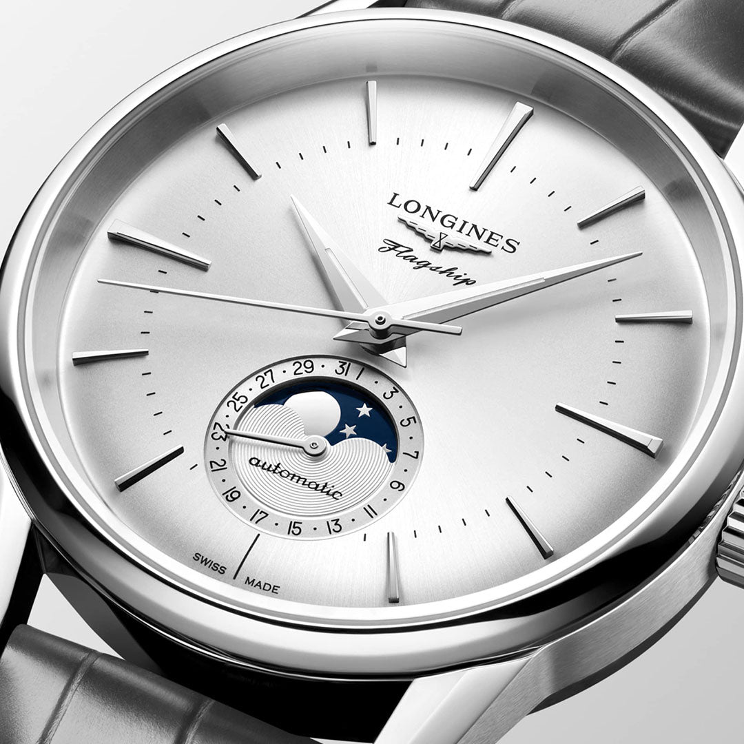 Close-up of the Longines Flagship Heritage Automatic 38.5mm Watch showcasing its silvered sunray dial. It includes slender hour markers, elegant hands, and a unique moon phase complication at the 6 o'clock position with a rotating disc featuring the moon and stars. The strap is metallic.