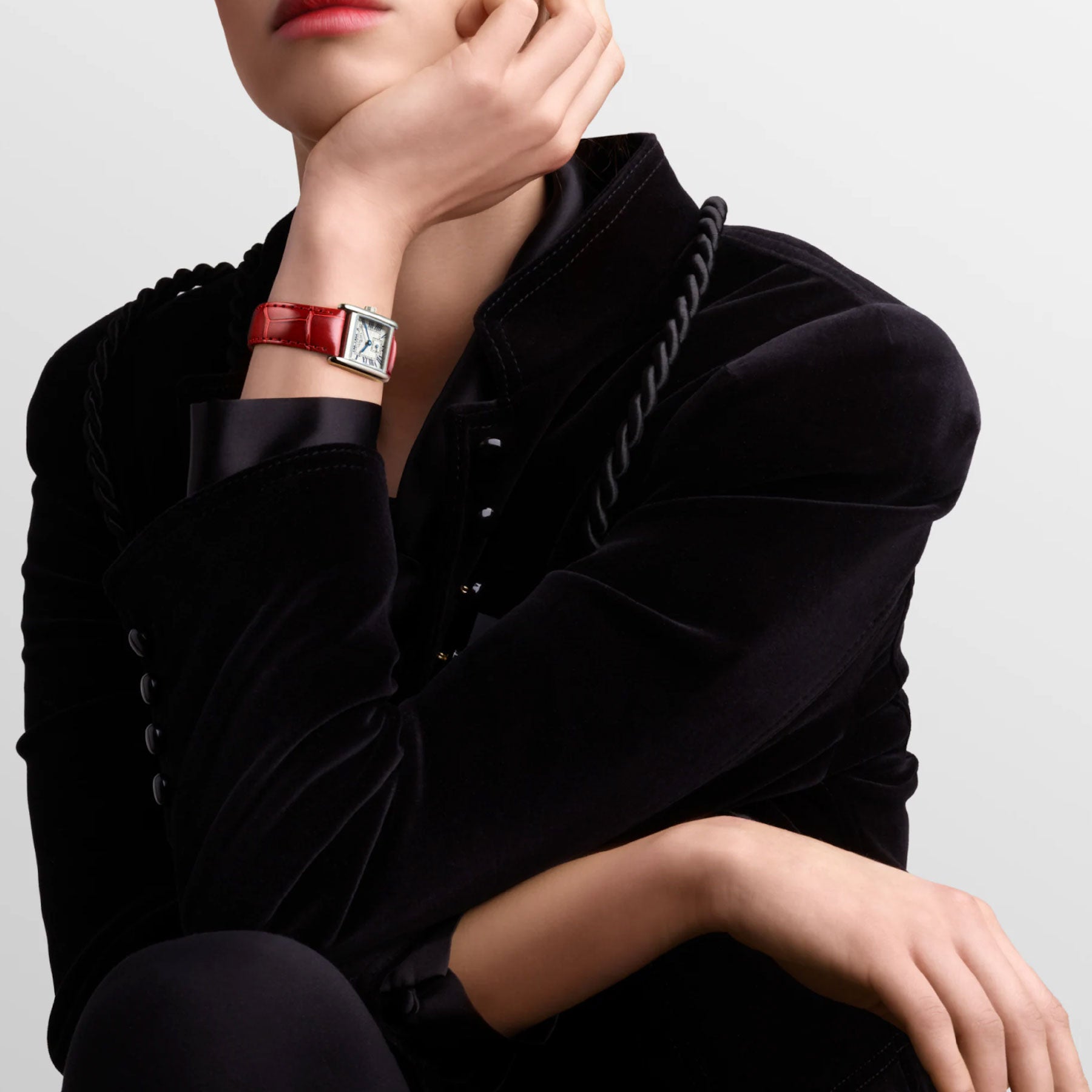 A person adorned in a black velvet jacket is seated, their chin gently resting on one hand while featuring the Longines Mini Dolcevita Quartz 21.5 x 29mm Watch. The red leather strap perfectly enhances the stainless steel case. The backdrop is a simple, light hue.