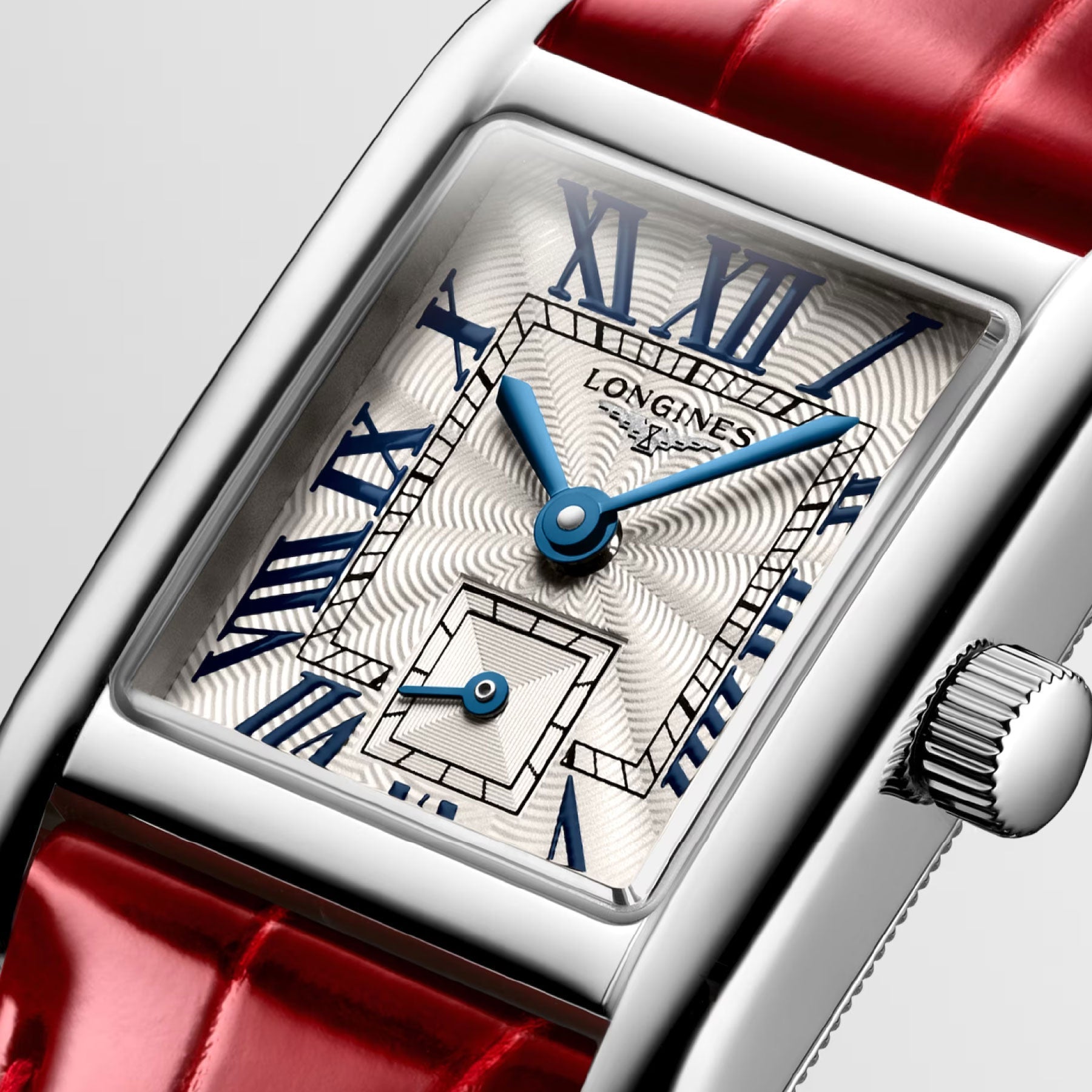 Close-up of the Longines Mini Dolcevita Quartz 21.5 x 29mm Watch, showcasing a rectangular stainless steel case with a patterned white face. The dial features blue Roman numerals, blue hands, and the brand name "Longines." It comes with a red leather strap and has a crown on the side.