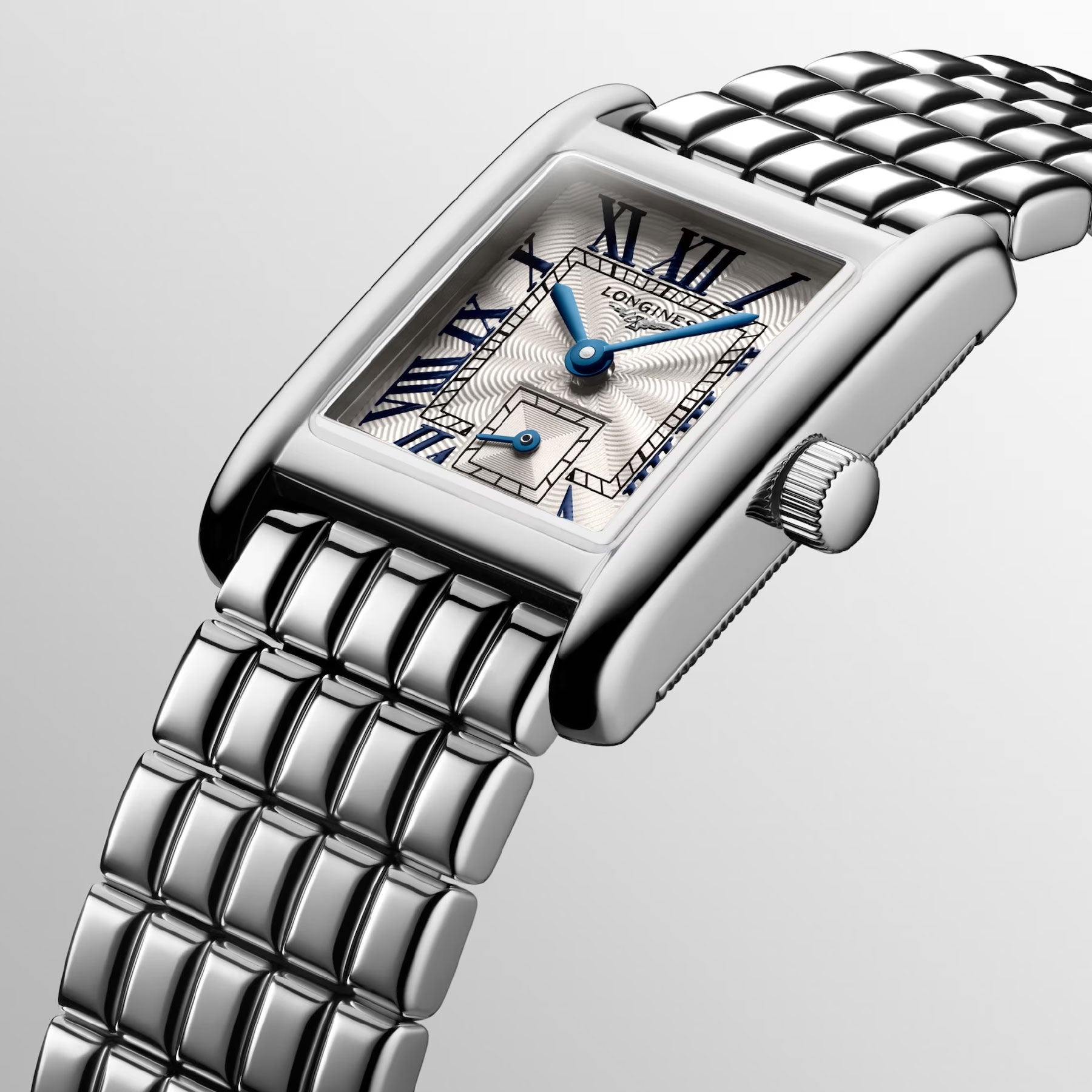 A close-up of a Longines Mini Dolcevita Quartz 21.5 x 29mm Watch showcases its luxury design with a stainless steel case, silver metal bracelet, and rectangular face. Featuring Roman numerals, blue hands, and intricate detailing on the dial, this timepiece offers quartz movement against a gradient gray background.