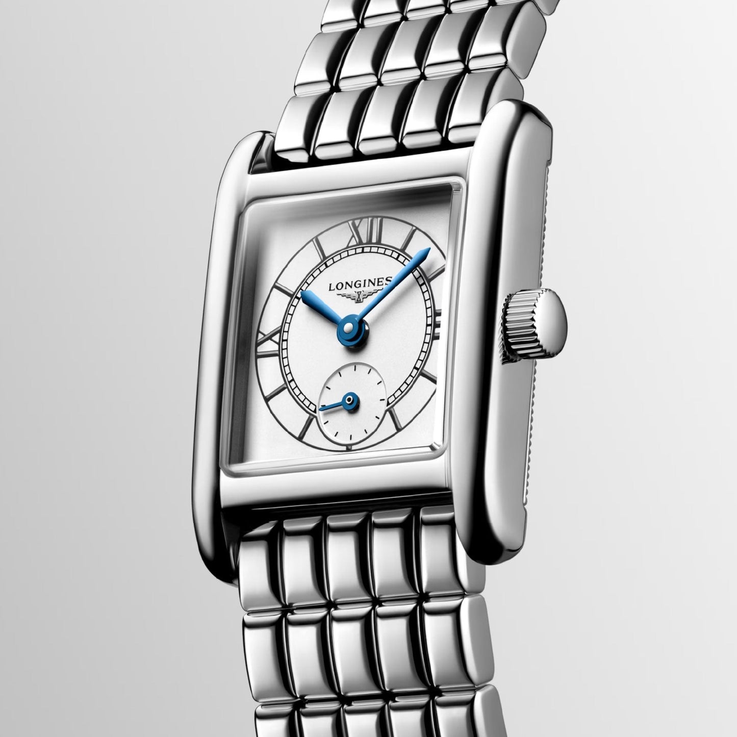 A close-up image of the Longines Mini Dolcevita Quartz 21.5 x 29mm watch highlights its silver wristwatch design with a rectangular face. This elegant timepiece showcases blue hands, a small subdial, and Roman numerals set against a white background. The watch features a polished metallic strap with a grid pattern and a ridged crown, emphasizing its stainless steel case from Longines.