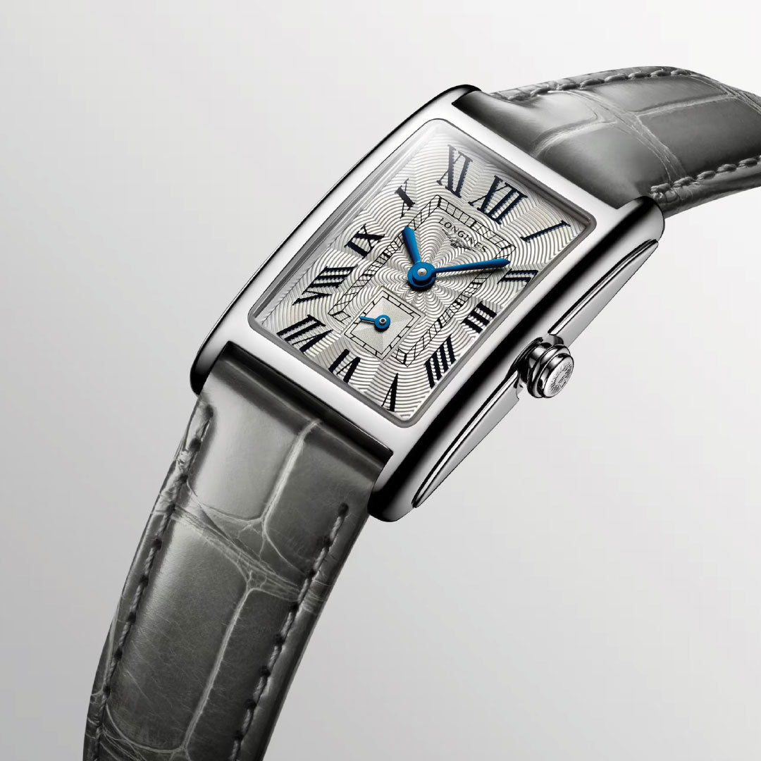 The Longines Dolcevita Quartz 20.8 x 32mm watch showcases modern elegance with a silver rectangular face, Roman numerals, and elegant blue hands on a textured dial. Its grey crocodile-patterned leather strap complements the light gray gradient background, enhancing its timeless charm.