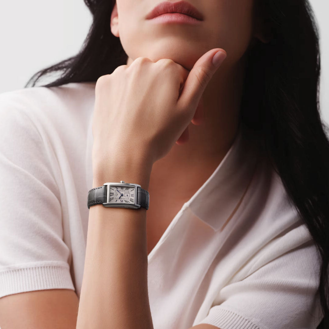 With dark hair, a person in a white shirt and wristwatch embodies modern elegance. The Longines Dolcevita Quartz 20.8 x 32mm Watch, featuring a rectangular face and black strap, is reminiscent of classic watchmaking and perfectly highlighted as their hand rests near their chin.