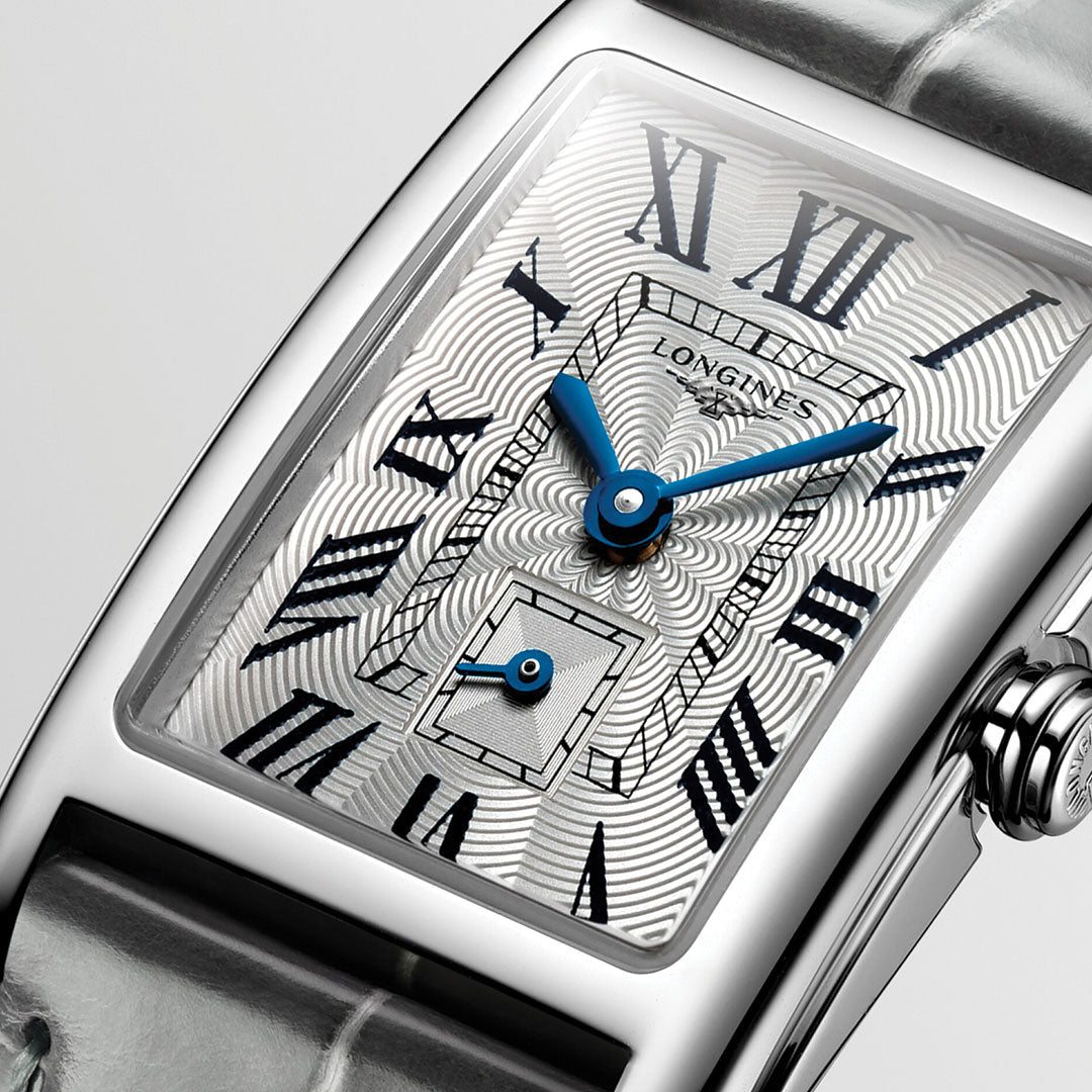 Close-up of the Longines Dolcevita Quartz watch (20.8 x 32mm) featuring a silver-toned face with blue hands, Roman numerals, and a sub-dial. The metallic case exudes elegance with intricate patterns and is paired with a glossy gray leather strap.