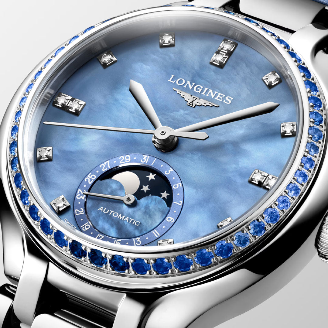 A close-up of the Longines Primaluna Moonphase Automatic 34mm watch shows its blue mother-of-pearl dial, diamond markers, sapphire-set bezel, moon phase subdial, and silver strap. The women's watch hands are set to 10:08.