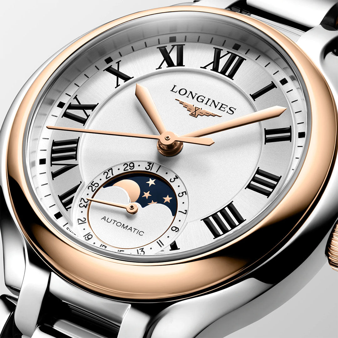 A close-up of the Longines Primaluna Moonphase Automatic 34mm Watch, featuring a silver and gold case, Roman numerals, gold hands, and a moon phase/date subdial with "AUTOMATIC" visible above it. Brand: Longines.