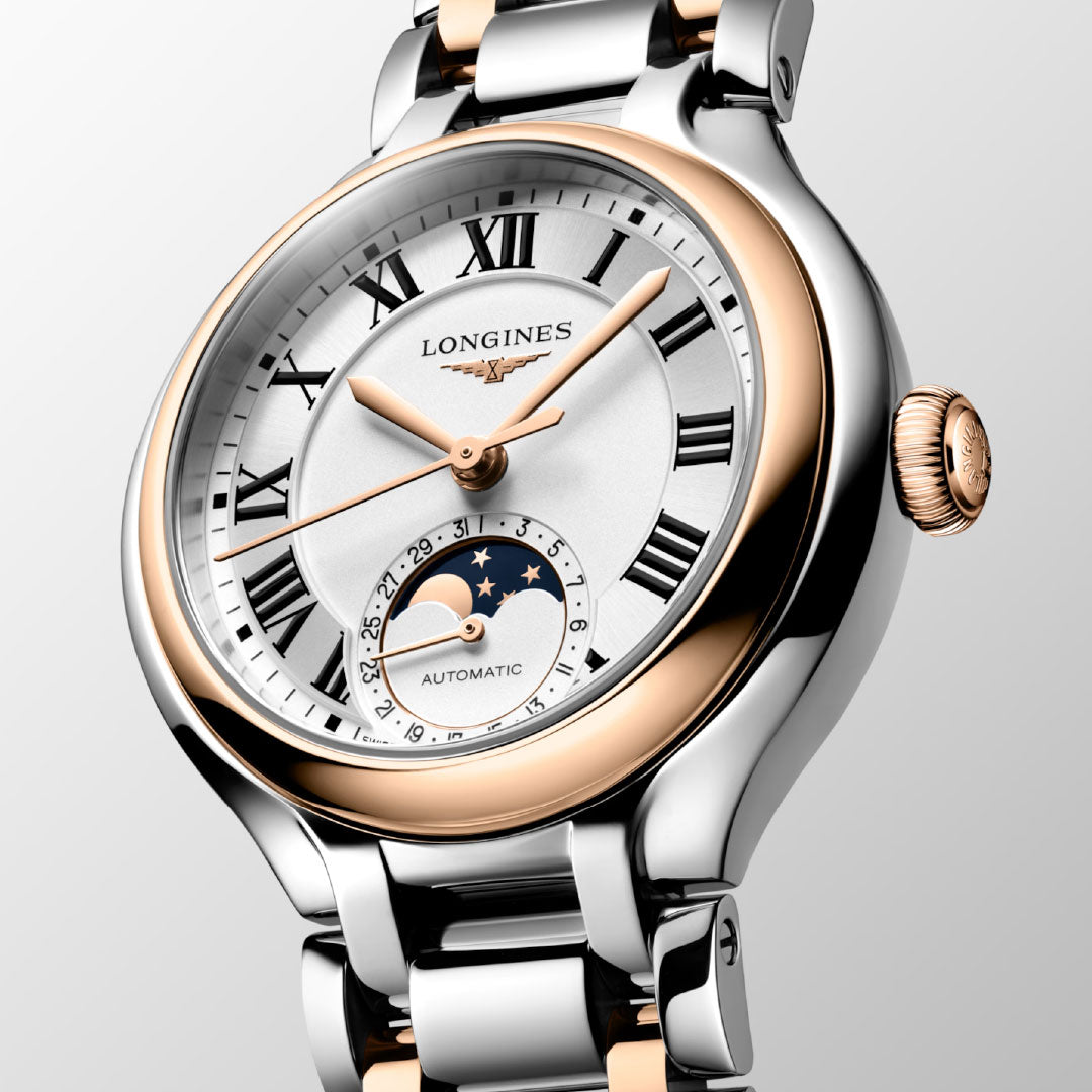 A close-up of the Longines Primaluna Moonphase Automatic 34mm women's watch, featuring a silver and rose gold band, Roman numerals, moon phase subdial on a white dial, and gold hands. The dial displays "automatic.