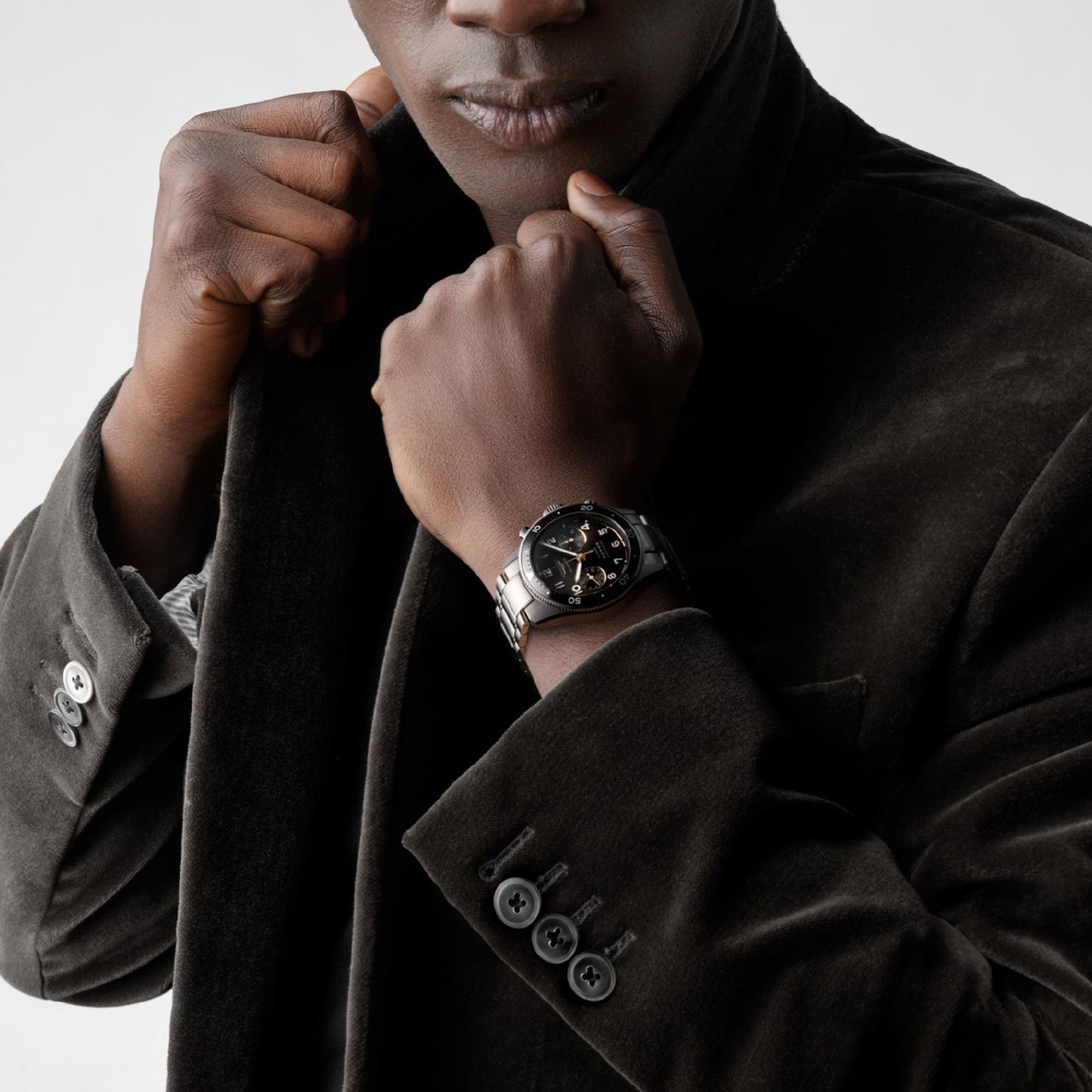 A person in a dark velvet jacket holds their collar with both hands, showcasing the elegant Longines Spirit Flyback Automatic Chronograph 42mm watch, a piece celebrated for its association with legendary pioneers. The plain, light background highlights the fine details of the outfit and the intricate design of the watch.