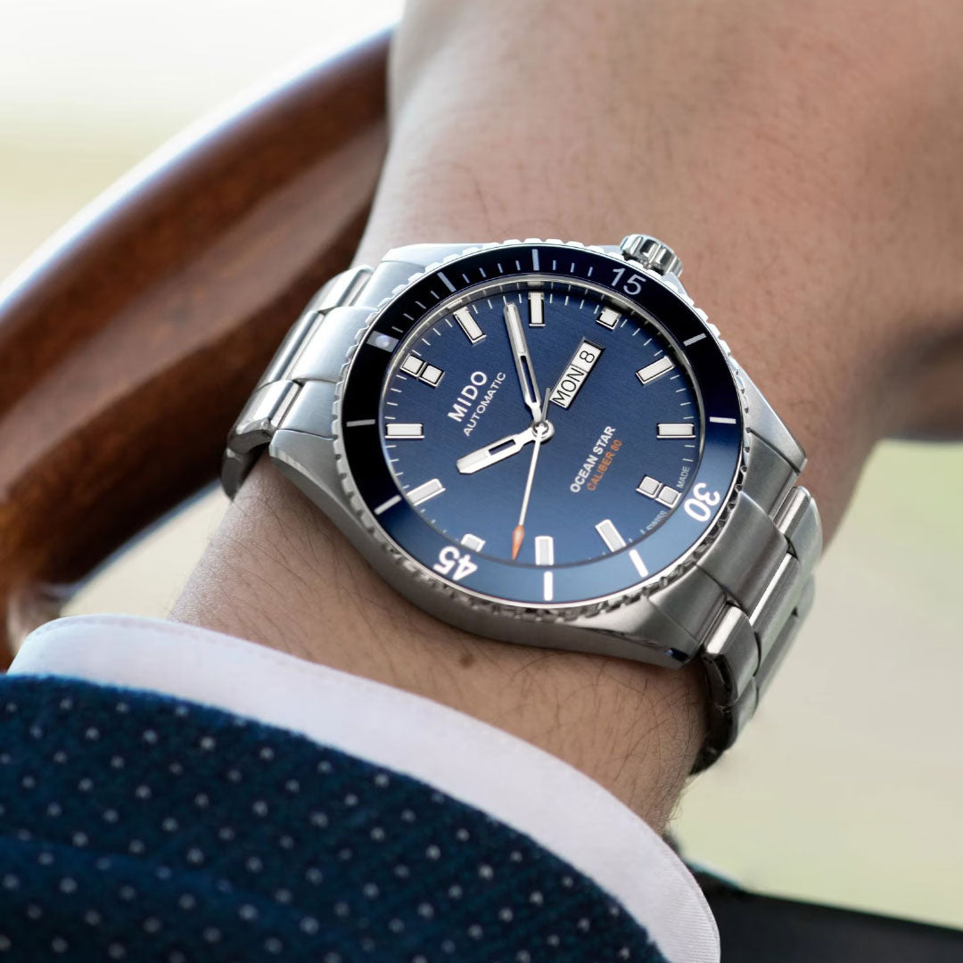 A person in a navy blue dotted suit wearing the MIDO Ocean Star 200 Automatic 42.5mm Watch with a blue dial and day-date display holds a wooden steering wheel.