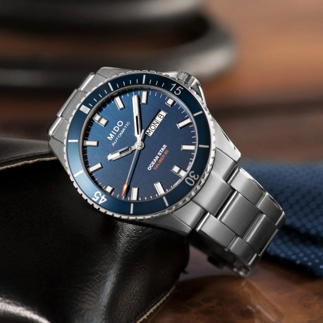 A MIDO Ocean Star 200 Automatic 42.5mm silver watch with a blue dial and silver bracelet rests on brown leather, with a blue tie and blurred objects in the background.