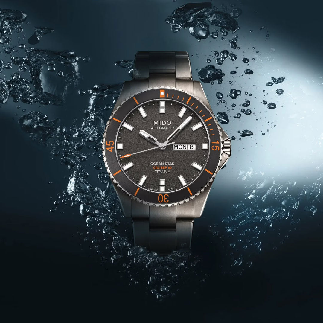 The MIDO Ocean Star 200 Automatic 42.5mm watch, featuring a titanium case and orange-accented bezel with a black strap, is highlighted in dramatic light with splashing water, showcasing its day-date display and water resistance.