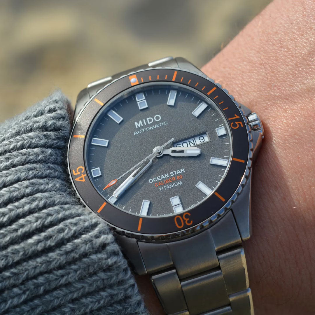 A close-up of a wrist wearing a MIDO Ocean Star 200 Automatic 42.5mm Watch with a grey dial, orange accents, and stainless steel band, displaying the day and date. The wearer has on a grey knit sweater.
