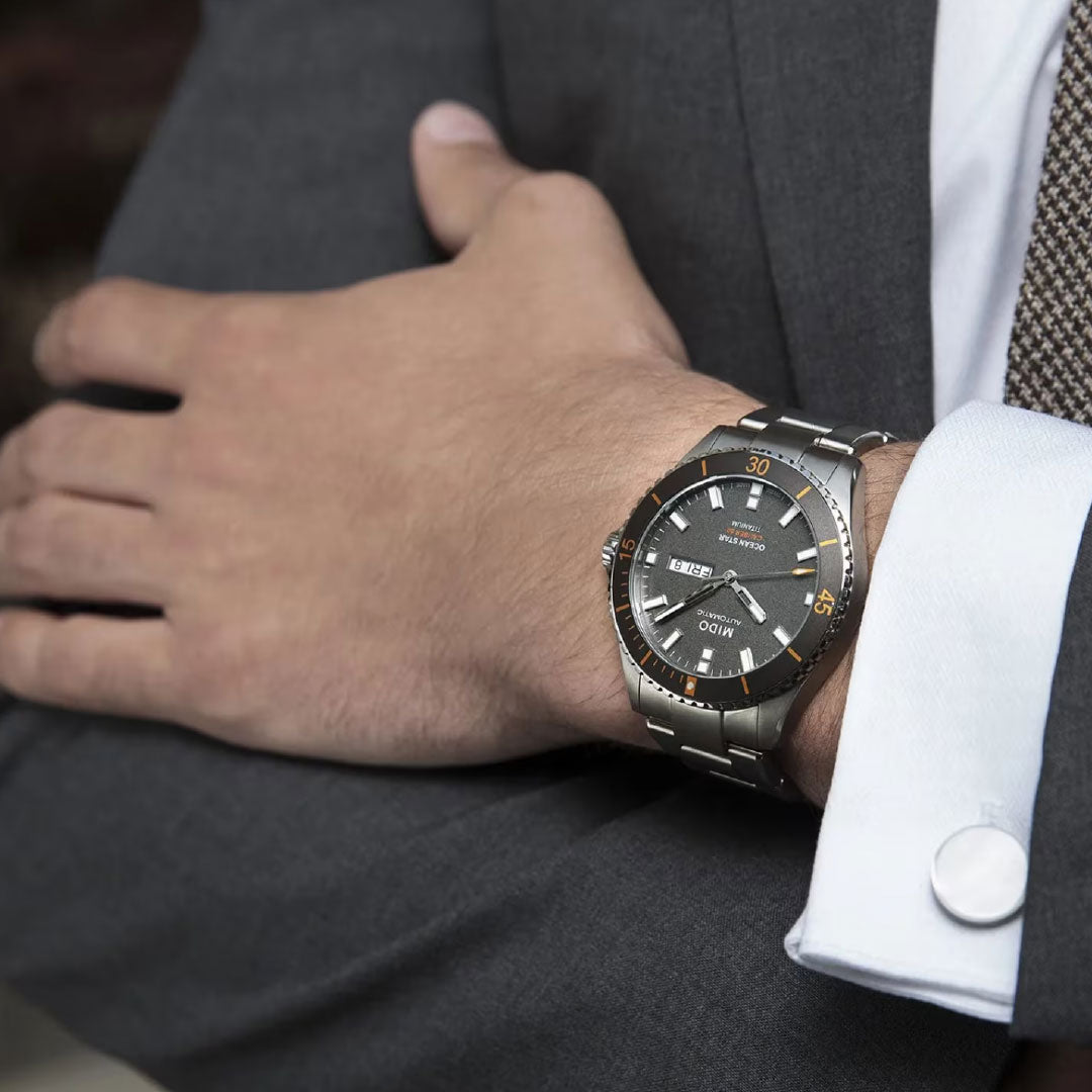 A person in a gray suit and patterned tie, arms crossed, wears a MIDO Ocean Star 200 Automatic 42.5mm Watch on their left wrist.