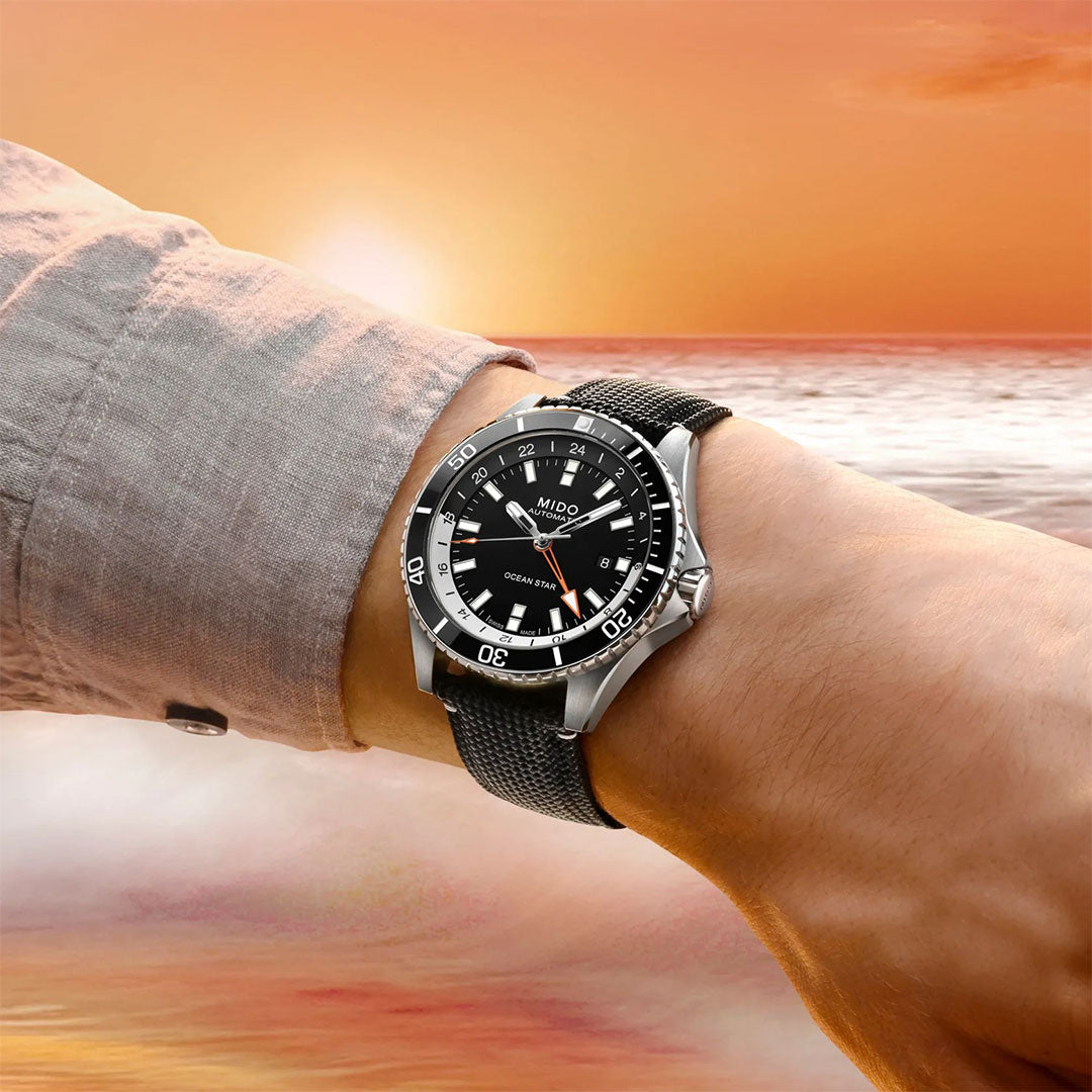 A person wearing the MIDO Ocean Star GMT Automatic 44mm Watch with a black dial and silver case stands by the ocean at sunset, with waves and an orange sky in the background.