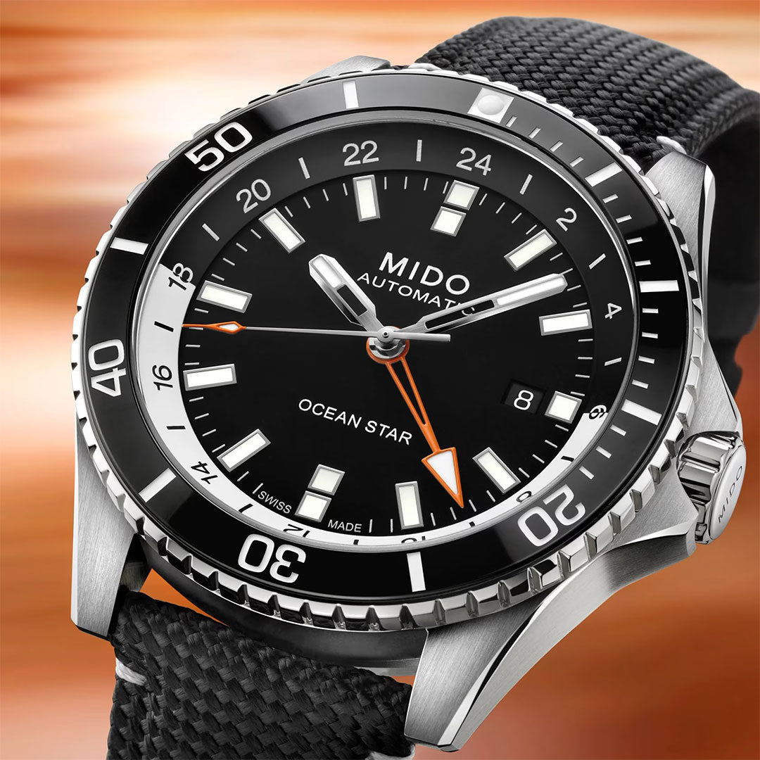 A close-up of the MIDO Ocean Star GMT Automatic 44mm watch features a black dial, white hour markers, date display, and a black textured strap, set against a blurred brown background.