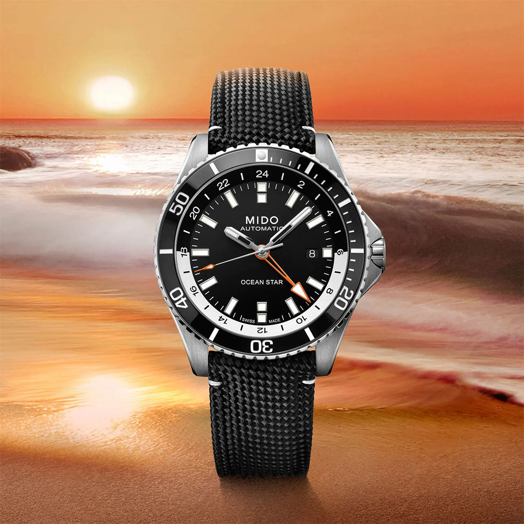 A MIDO Ocean Star GMT Automatic 44mm Watch by MIDO, featuring a black strap, is displayed in front of a beach at sunset with waves and the sun visible against an orange sky background.