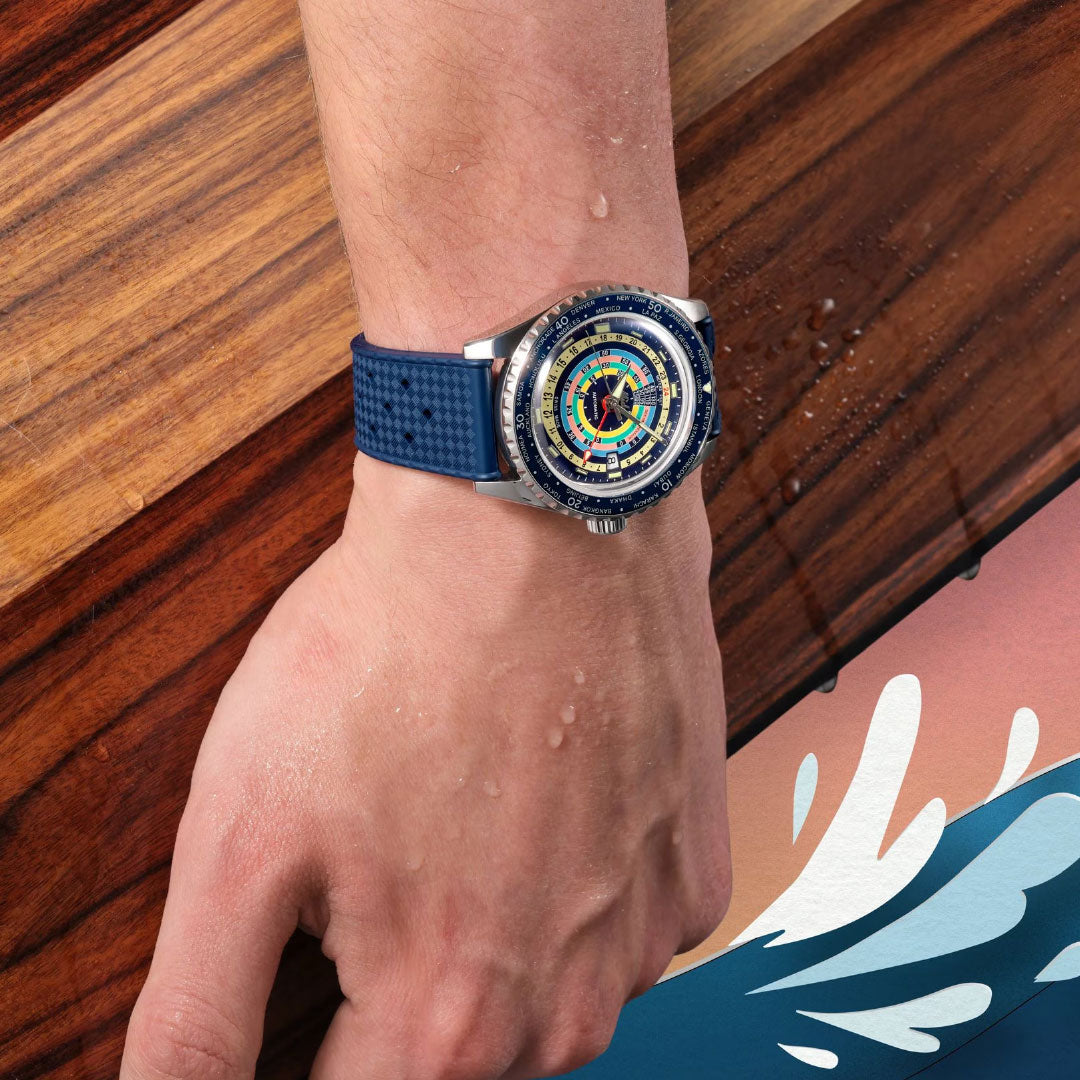 A close-up shows a person's left wrist wearing the MIDO Ocean Star Decompression World Timer Automatic 40.5mm Watch with a blue strap, water droplets on the hand, and a wooden wave graphic in the background.