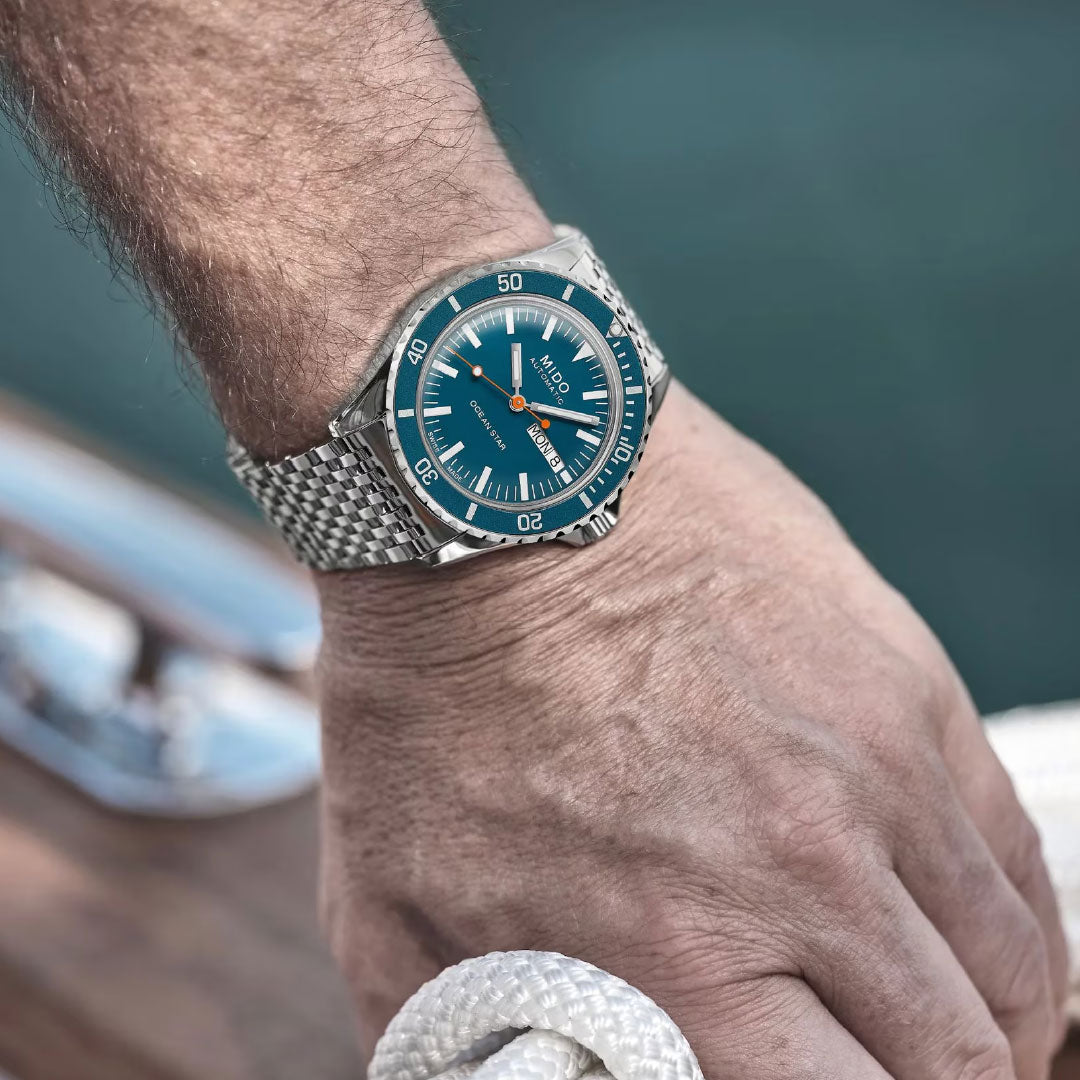 A close-up of a person's wrist wearing a MIDO Ocean Star Tribute Automatic 40.5mm Watch with a blue face and mesh bracelet, holding a white rope, with water and a boat deck blurred in the background.