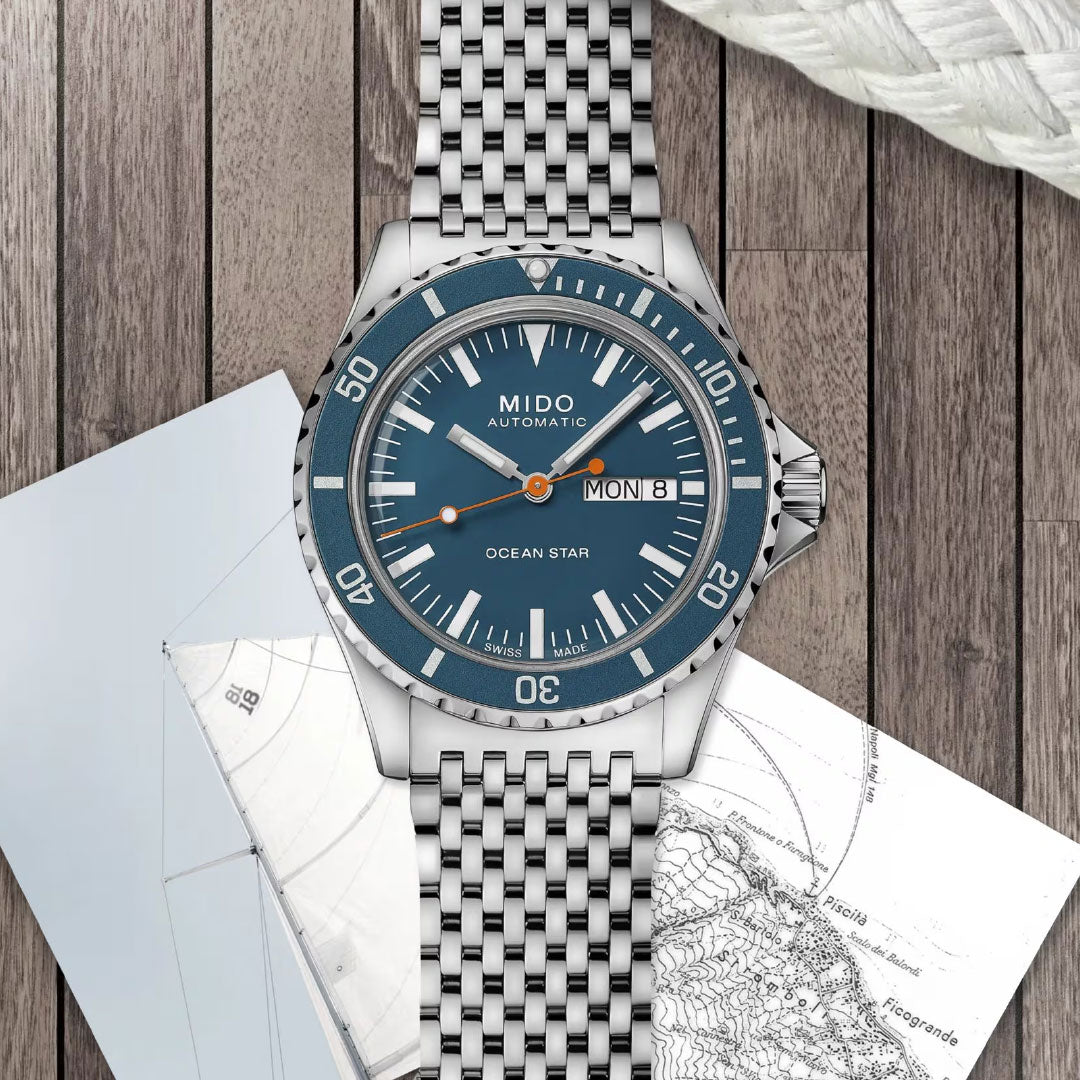 The MIDO Ocean Star Tribute Automatic 40.5mm Watch features a blue dial and day-date display, powered by the Caliber 80 movement. Set on wooden planks beside nautical charts and coiled rope, it's made for maritime adventure.
