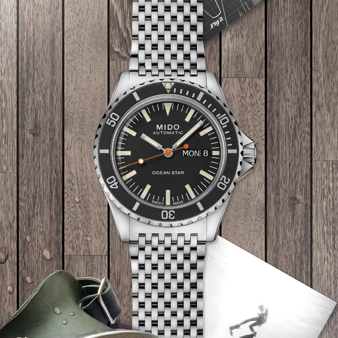 A MIDO Ocean Star Tribute Automatic 40.5mm watch with a black dial and day-date display rests on a wooden surface, surrounded by black-and-white photos and a diving mask.