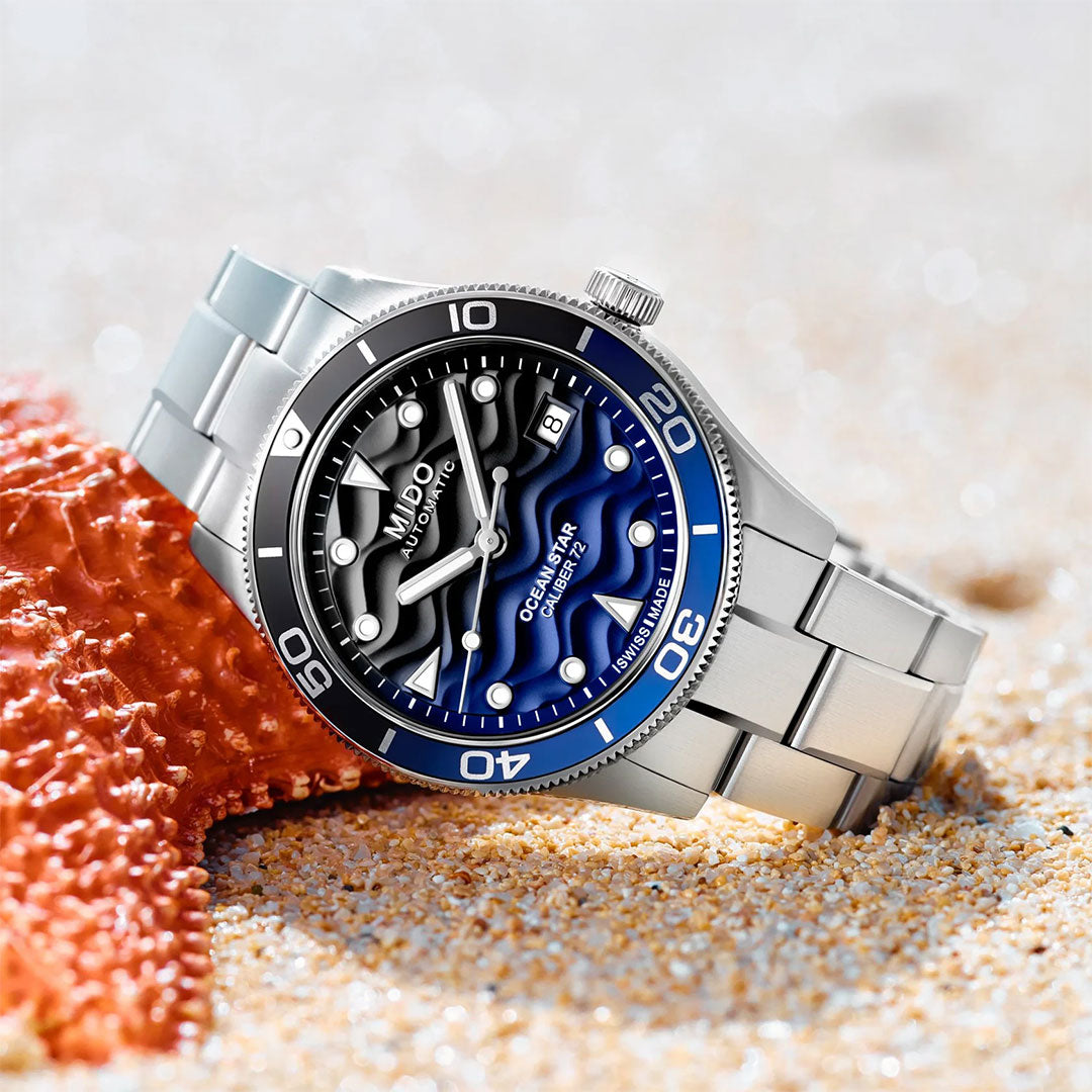 The MIDO Ocean Star 39mm Automatic watch, featuring a blue and black dial and water-resistant face, rests on sandy beach beside an orange starfish, showing the time and date.