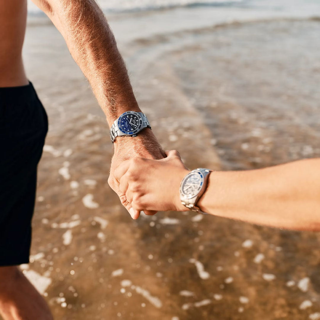 Two people stand hand in hand in shallow beach water, sunlight reflecting off their arms as they wear stylish, water-resistant MIDO Ocean Star 39mm Automatic watches, with gentle ocean waves shimmering in the background.