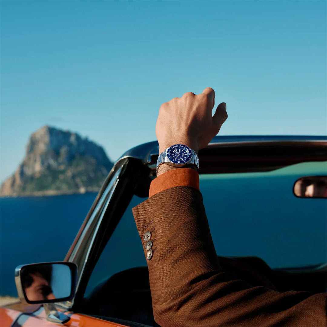 A person in a brown suit raises their left arm out of a car window, displaying the MIDO Ocean Star 200 Automatic 41mm Watch with its blue face. Behind them, the ocean and a rocky island shine beneath the clear blue sky.