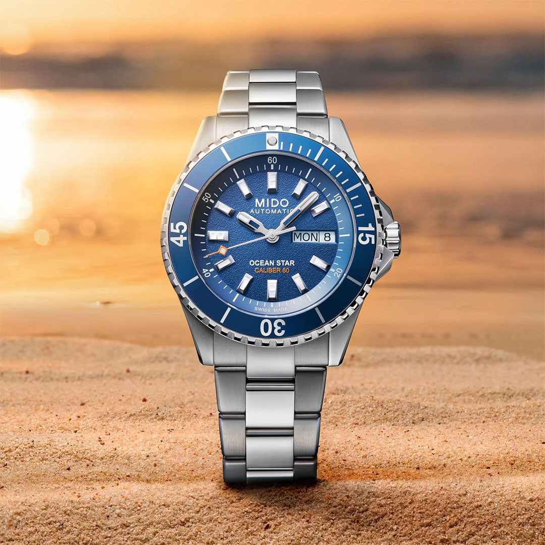 The MIDO Ocean Star 200 Automatic 41mm Watch is showcased upright on sandy beach, its silver case and blue dial/bezel highlighted by a blurred ocean sunset in the background.