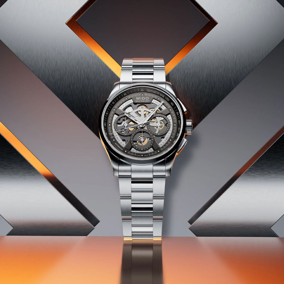 The MIDO Multifort Skeleton Chronograph Automatic 43mm watch, featuring a silver tone and metal bracelet, is showcased upright against a geometric metallic backdrop with orange lighting accents.