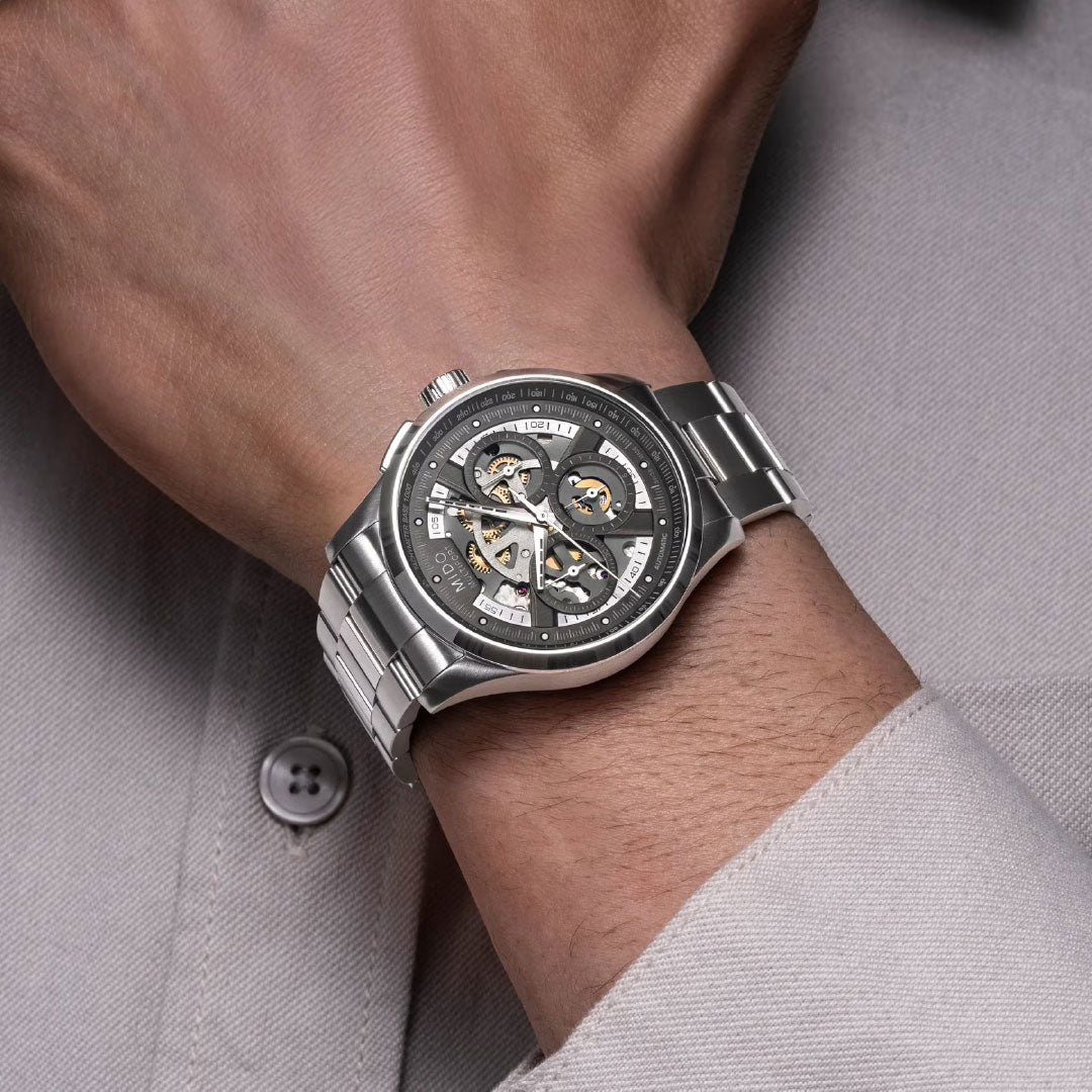 A person wearing the MIDO Multifort Skeleton Chronograph Automatic 43mm Watch, featuring a metallic band and exposed gears, on their wrist over a light gray button-up shirt.