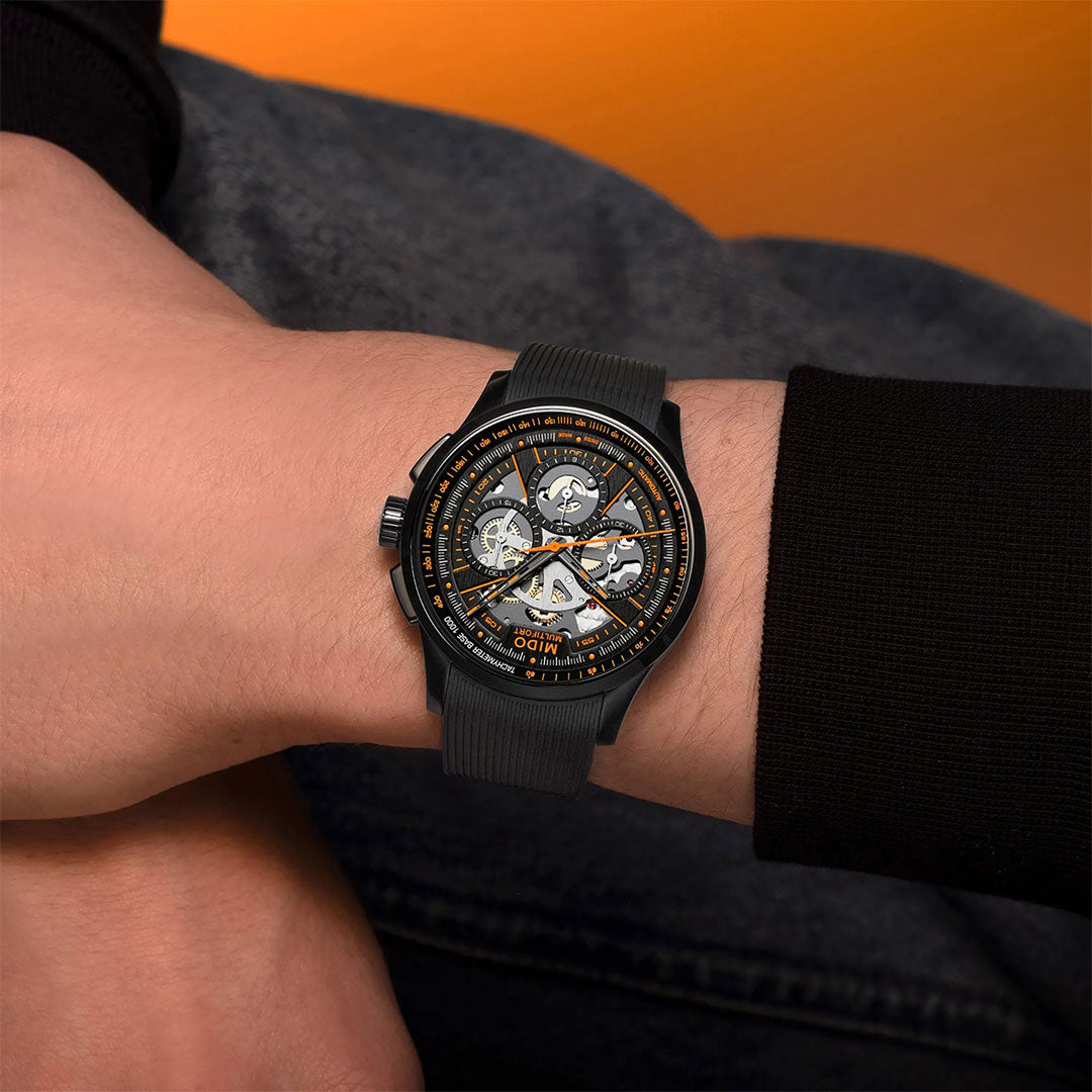 A person wearing the MIDO Multifort Skeleton Chronograph Automatic 43mm Watch, featuring orange accents, a skeleton dial, and a black strap, rests their wrist on their leg against an orange background.