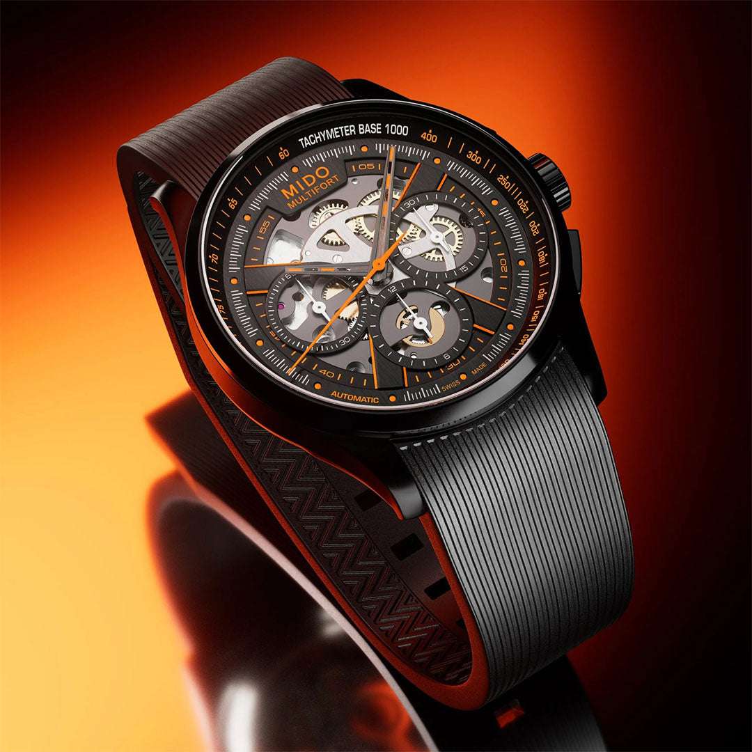 A close-up of the MIDO Multifort Skeleton Chronograph Automatic 43mm Watch, featuring a skeleton dial with visible gears, Swiss automatic movement, black rubber strap, and tachymeter bezel against a glowing orange background.