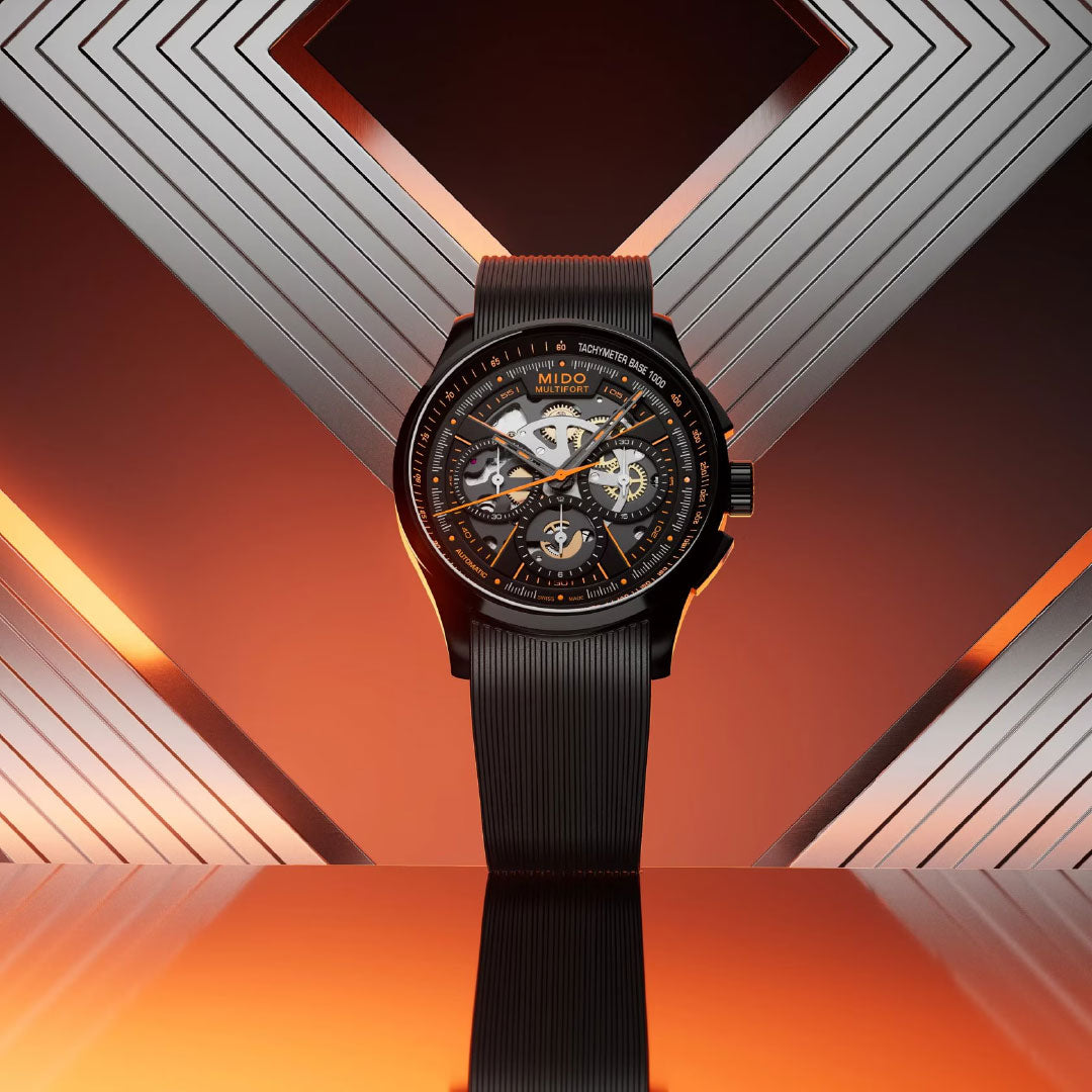 The MIDO Multifort Skeleton Chronograph Automatic 43mm watch is showcased upright against a metallic geometric background with orange lighting, highlighting its intricate skeleton dial and modern design.